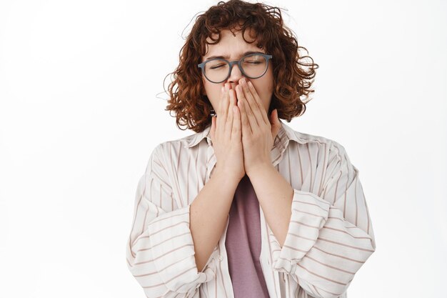 Cute young woman with curly hair and glasses, yawning, covering opened mouth while yawn sleepy, feel tires, sneezing from allergy, standing against white background