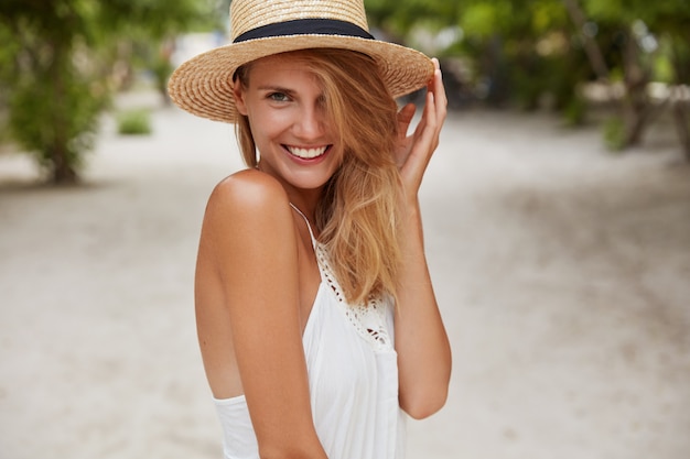 Free photo cute young woman with shining smile, has tanned healthy skin and appealing look, enjoys summer rest in paradise place, wears straw hat, smiles pleasantly. people, beauty and seasonal rest concept