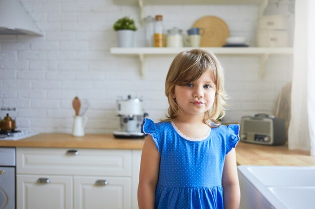 Cuteness, sweet tender age and interior design concept. Portrait of cute adorable female child in blue dress pouting lips, having displeased facial expression, posing in kitchen, 