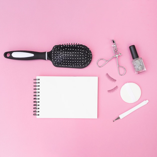 Cuticle; hair brush; sponge; fake eyelashes; eyelash curler; nail polish bottle with blank spiral notepad on pink backdrop