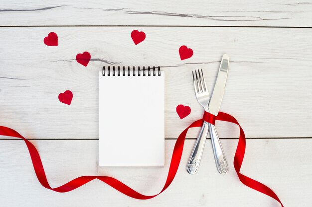 Cutlery with ribbon near ornament hearts and notebook