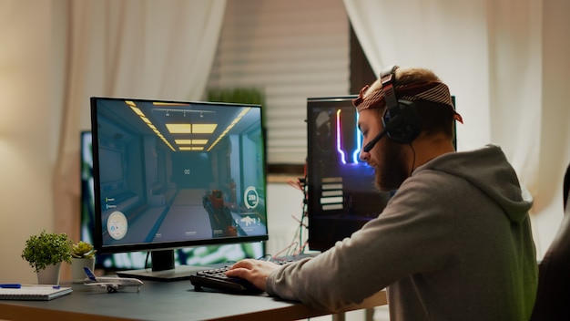 Cyber sport gamer wearing headphones playing first person shooter videogame participating in eSports tournament perforing at RGB prowerful personal computer. Pro cyber streaming gaming championship