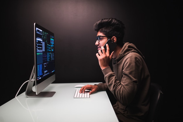 Cybercrime, hacking and technology concept. male hacker with smartphone and coding on computers screen in dark room