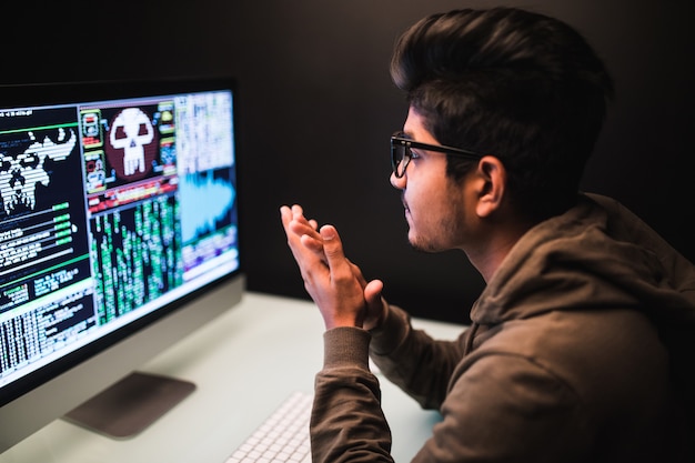 Cybercrime, hacking and technology concept - Young male hacker with smartphone and coding on computers screen in dark room