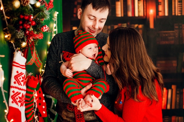 Dad holds kid in stripped suit standing before a Christmas tree while mother plays with her 