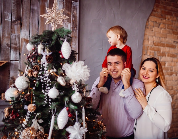 Dad holds little daughter  on his neck posing with mom before a rich Christmas tree