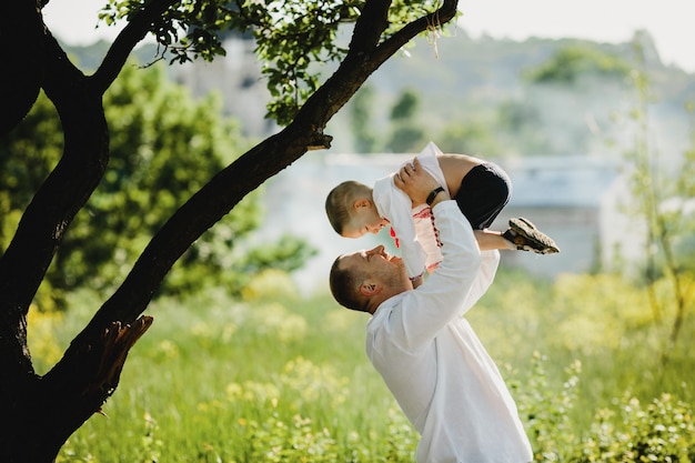 Free photo dad holds little son in embroidered shirt in his arms standing under green tree