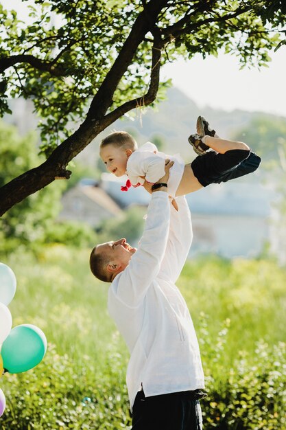 Dad holds little son in embroidered shirt in his arms standing under green tree