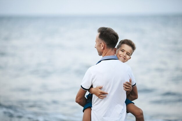 Dad is holding son on the hands and child is looking straight and smiles on the seashore