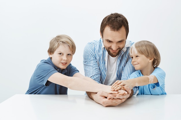 Dad and sons not only family but team. Portrait of happy good-looking siblings and father holding hands while sitting at table, smiling broadly