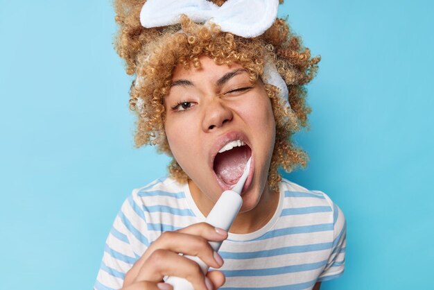 Ежедневная утренняя рутина и концепция привычек чистки Молодая женщина-модель с вьющимися светлыми волосами чистит зубы электрической зубной щеткой, держит зубы здоровыми, изолированными на синем фоне.