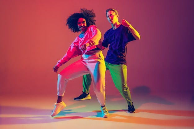 Dance time. Stylish men and woman dancing hip-hop in bright clothes on green background at dance hall in neon light. 