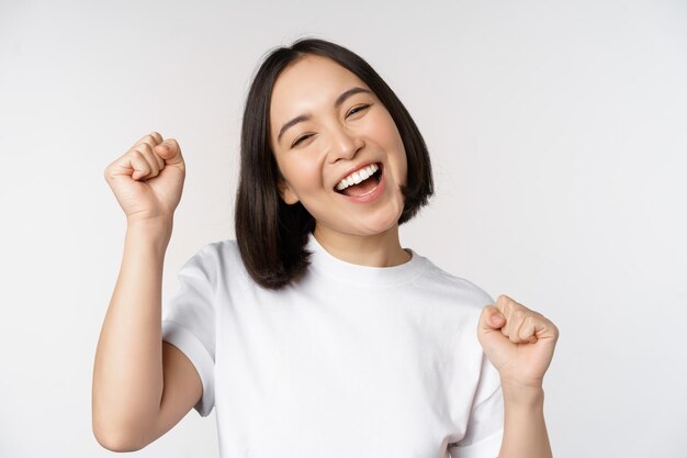 Dancing asian girl celebrating feeling happy and upbeat smiling broadly standing over studio white background