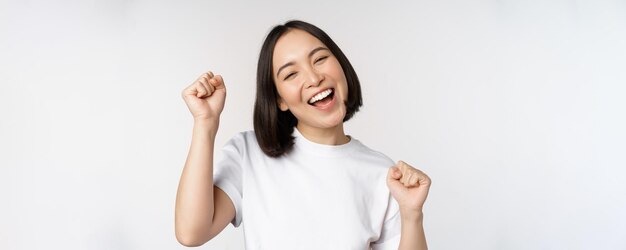 スタジオの白い背景の上に広く立って幸せで明るい笑顔を祝って踊るアジアの女の子