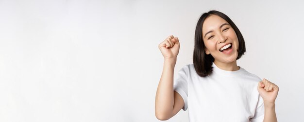 스튜디오 흰색 배경 위에 활짝 서서 행복하고 낙관적인 미소를 지으며 축하하는 춤추는 아시아 소녀