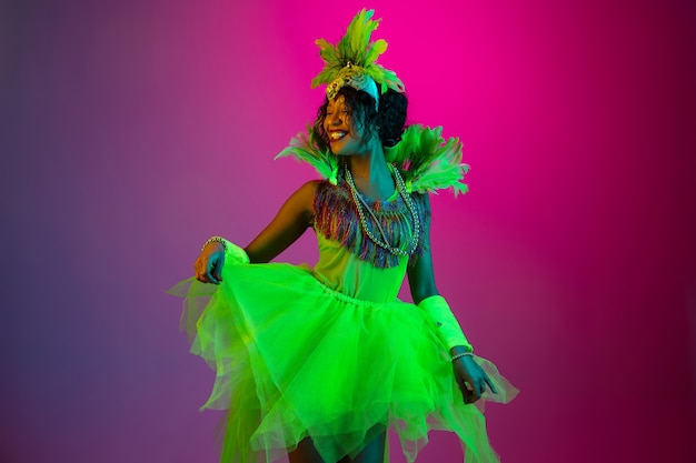 Dancing. Beautiful young woman in carnival, stylish masquerade costume with feathers dancing on gradient background in neon. Concept of holidays celebration, festive time, dance, party, having fun.