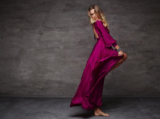 Dancing blond girl in long red dress on grey background