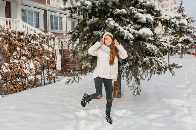 Dancing caucasian woman in cute knitted hat. Outdoor full-length portrait of blissful long-haired lady in ripped pants fooling around near snowy tree..