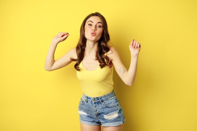 Dancing happy summer girl having fun, raising hands up and rejoicing, standing in tank top and shorts against yellow background.