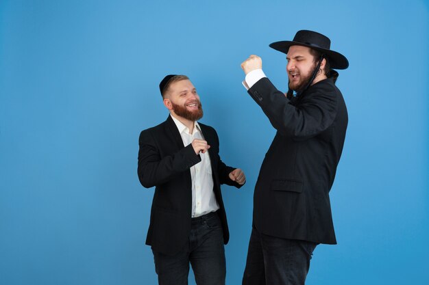 Dancing, having fun. Portrait of a young orthodox jewish men isolated on blue  wall. Purim, business, festival, holiday, celebration Pesach or Passover, judaism, religion concept.