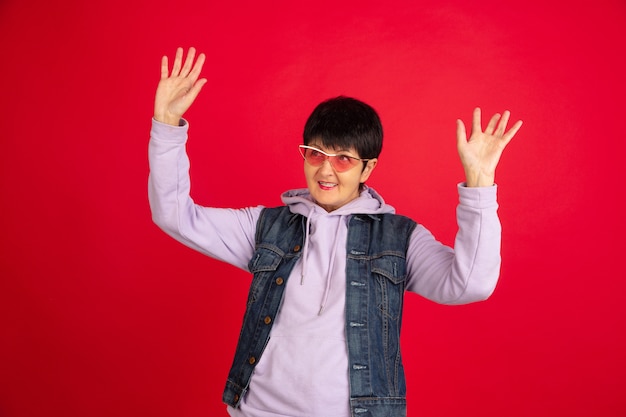 Dancing. Portrait of senior woman in stylish outfit, attire isolated on red studio background. Tech and joyful elderly lifestyle concept. Trendy colors, forever youth. Copyspace for your ad.