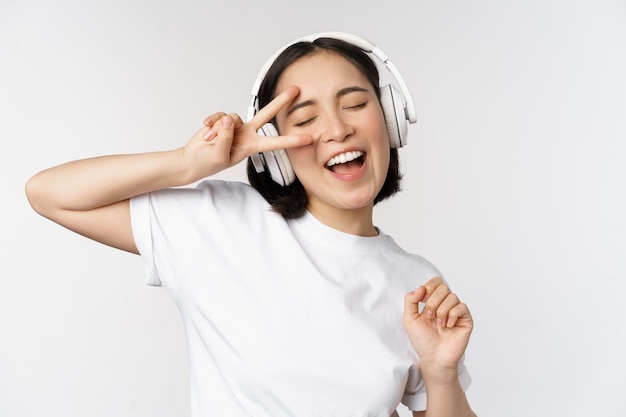 Dancing and singing asian woman listening music in headphones standing in earphones against white background Copy space