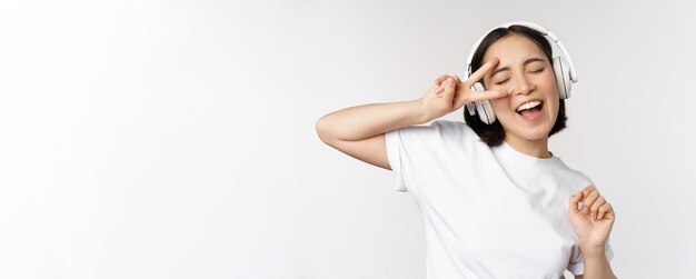 白い背景のイヤホンで立っているヘッドフォンで音楽を聴いて踊って歌うアジアの女性