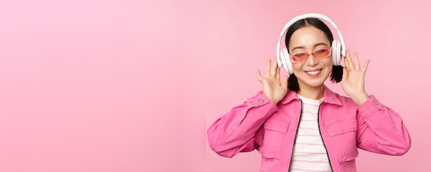 Dancing stylish asian girl listening music in headphones posing against pink background