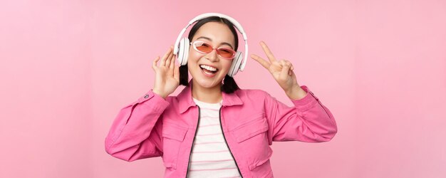 Dancing stylish asian girl listening music in headphones posing against pink background