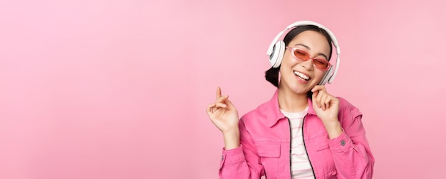 Танцующая стильная азиатская девушка слушает музыку в наушниках, позируя на розовом фоне