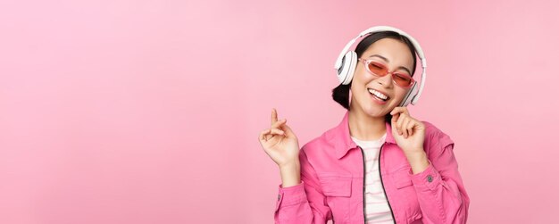 Dancing stylish asian girl listening music in headphones posing against pink background