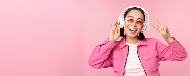 Dancing stylish asian girl listening music in headphones posing against pink background