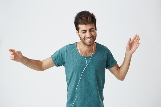 Dancing stylish bearded student in casual clothes. Waving his hands, enjoying his favourite songs. Guy wearing white earphones and smiling