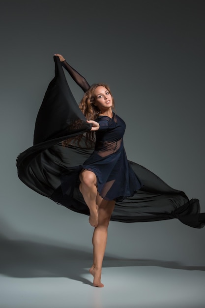 Танцующая женщина в черном платье. Современный современный танец на сером фоне. Фитнес, модель на растяжку