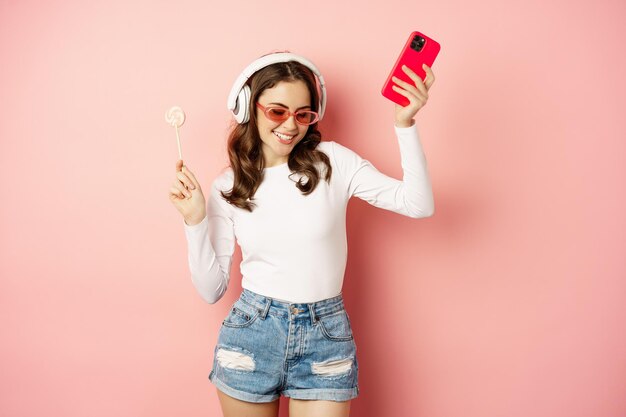 Танцующая женщина в солнечных очках, слушает музыку в наушниках, держит леденец на палочке и смартфон, смеется и счастливо улыбается, стоя на розовом фоне.