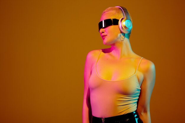 Dancing. Young caucasian woman on brown wall in neon light. Beautiful female model with fashionable, trendy eyewear. Human emotions, facial expression, sales, ad concept. Freak's culture.