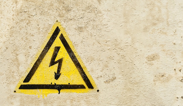 高電圧電気の危険兆候。古い灰色のひびの入った背景に稲妻と黄色の三角形のハザードサイン。コピースペースのクローズアップ