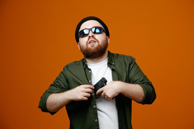 Dangerous looking bearded man wearing black glasses and hat holding a gun looking at camera boldly standing over orange background