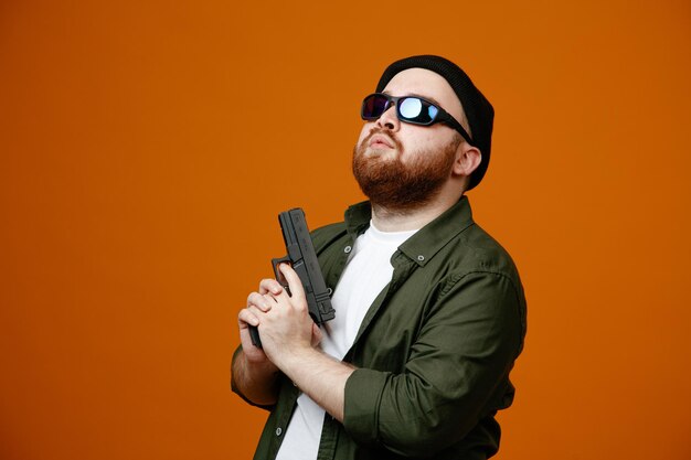 dangerous looking bearded man wearing black glasses and hat holding a gun looking confident with serious expression standing over orange background