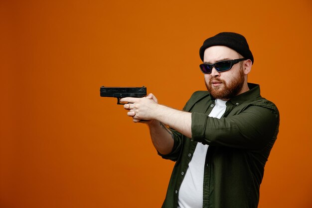 dangerous looking bearded man wearing black glasses and hat holding a gun pointing it at something looking aside standing over orange background