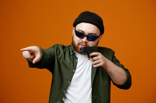 Dangerous looking bearded man wearing black glasses and hat holding a gun pointing with index finger at something standing over orange background