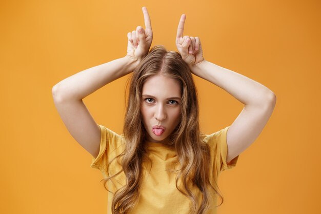 Daring and cool emotive attractive female student with wavy fair hair and little scars showing tongue rebellious looking from under forehead holding index fingers on head being stubborn. Copy space