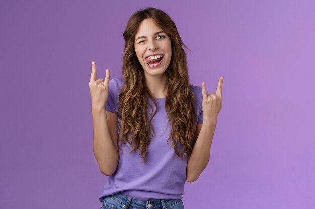 Daring sassy funny attractive woman having fun enjoy awesome party arrange music concert gathering show rock-n-roll heavy metal sign joyful stick tongue wink cheeky purple background. Lifestyle.