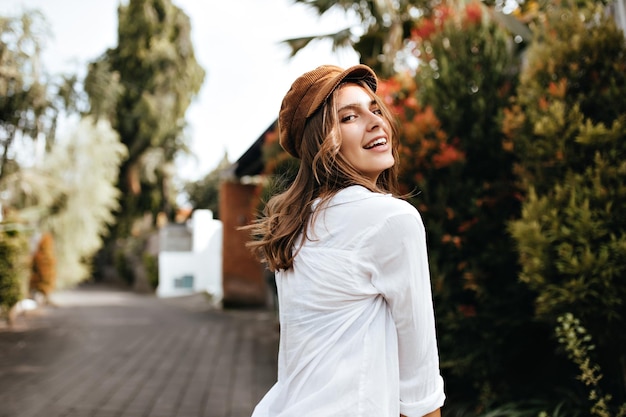 Free photo dark blonde cheerful girl in brown cap and white cotton blouse walks along path in park