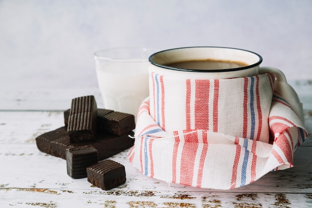 Foto gratuita barretta di cioccolato fondente; latte e tazza da caffè avvolti con tovagliolo sul tavolo di legno