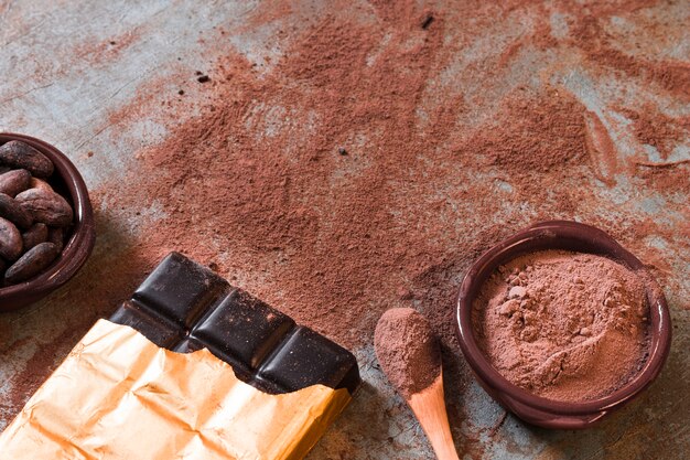 素朴な背景に散らばったココアパウダーと豆のボウルを持つダークチョコレートバー