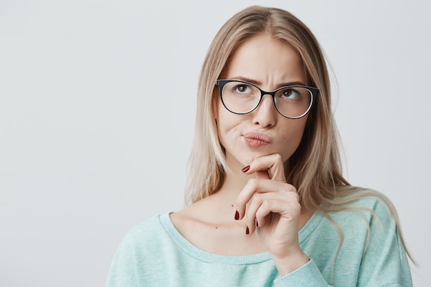 Dark-eyed blonde lovely young female with spectacles looks aside as has thoughtful expression and being concentraed on pleasant thoughts, dreams about something pleasant, has appealing appearance.