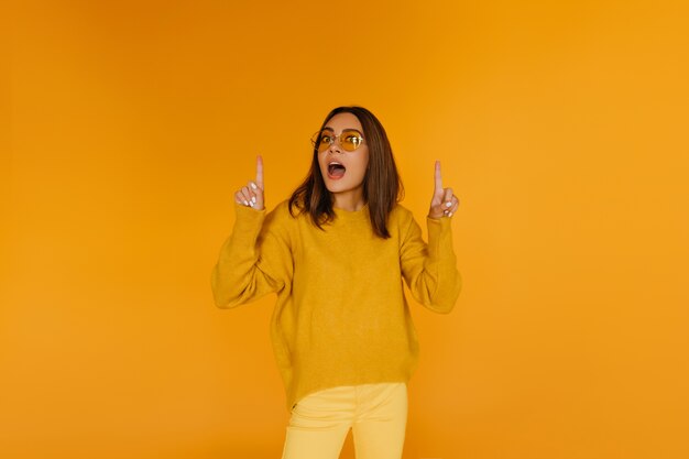 Dark-haired beautiful girl in glasses posing on yellow wall. Trendy european woman expressing amazement.