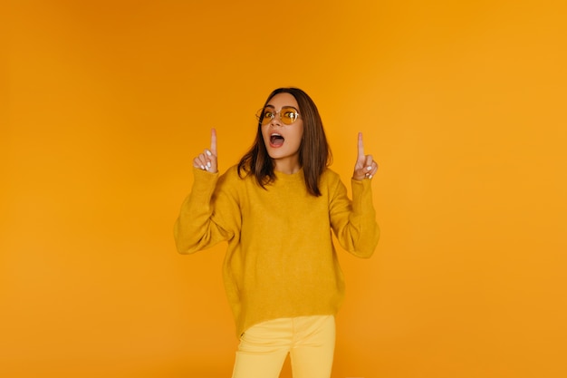 Dark-haired beautiful girl in glasses posing on yellow wall. Trendy european woman expressing amazement.