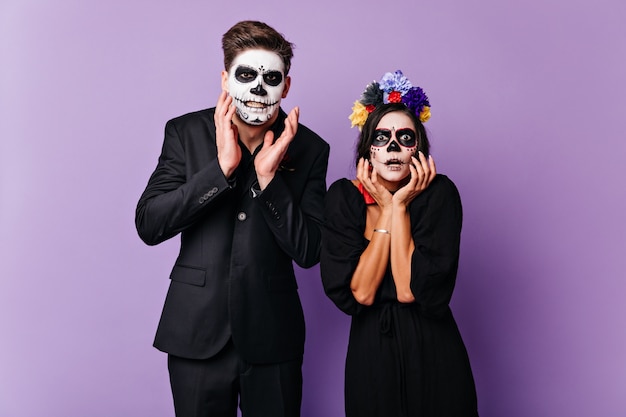 Dark-haired boy and girl in black outfit with Halloween make-up scared and surprised gestures